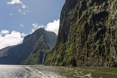Milford Sound 'daki sulardan yükselen bereketli yağmur ormanlarıyla kaplı devasa uçurumlar. Bir yüzeye bir şelale düşer ve küçük bir tekne Yeni Zelanda 'daki dramatik fiyorda ölçek sağlar..
