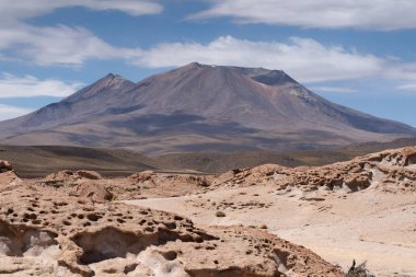 Zirvede sarı sülfür yatakları ve ön planda aşınmış kayalar bulunan Bolivya sınırındaki aktif Ollague Volkanı manzarası.