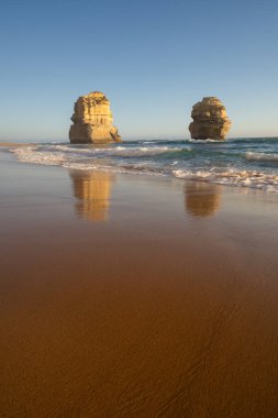 Islak kumda yansıyan iki belirgin deniz yığınının dikey, alçak açılı plaj manzarası, öğleden sonra güneşinin altında beyaz dalgalar tabanlarında kırılıyor..