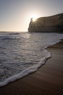 Düşük açılı, dikey bir sahil manzarası. Güneş, yüksek bir kumtaşı kayalık yüzeyinin gölgesinde parlıyor. Ön plandaki yansıtıcı nemli kumu yıkayan bir dalganın beyaz köpüğü..
