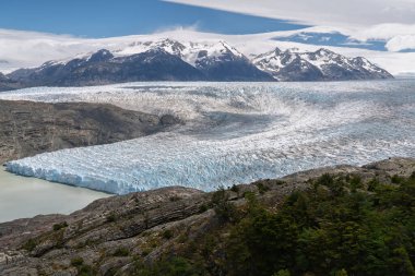Parlak bir gökyüzü altında, ihtişamlı, karla kaplı dağlarla desteklenen, Grey Gölü 'ne akan devasa, çatlamış buzullu buz tarlasının geniş, yüksek açılı görüntüsü..