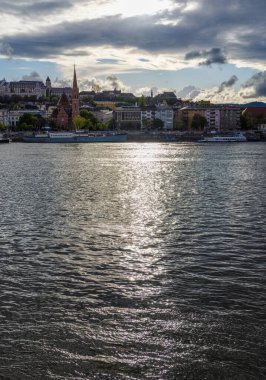 Scenic cityscape view across a calm river at sunset with sunlight reflecting off the water. Historic buildings and a church spire rise on the opposite bank under dramatic clouds
