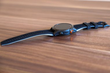 Parlak ahşap bir yüzeyde dinlenen parlak siyah bir akıllı. Modern tasarım, pürüzsüz kayış ve turuncu düğmeli metal kaplama günlük aksesuarlarda teknoloji ve stili sembolize ediyor.