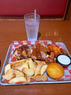 İçi füme et, tavuk kanadı, kalın kesilmiş patates kızartması, mısır ekmeği, daldırma sosu ve köpüklü bir içecekle dolu bir tepside servis edilen doyurucu bir barbekü yemeğine yakın plan..
