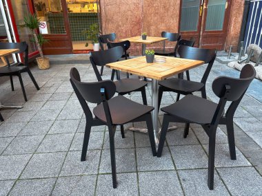 Boş açık kafe koltuklar, ahşap masalar ve taştan yapılmış teraslarda modern siyah sandalyeler. Sakin atmosfer ve minimalist mobilya tasarımı boş zaman ve modern yaşam tarzını çağrıştırıyor.