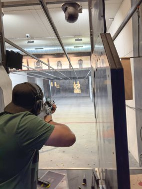 Kapalı alanda atış poligonuna nişan almış, hedeflere odaklanmış biri. Güvenlik teçhizatı, koruyucu bariyerler ve yerde boş kovanlarla kontrol edilen bir ortam..