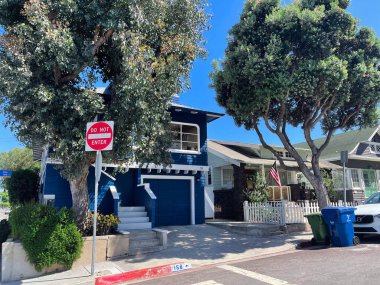 Los Angeles, ABD - 28 Ağustos 2024: Parlak güneşli banliyö sokak sahnesi garajı, beyaz süslemesi ve olgun ağaçları olan mavi ahşap bir ev