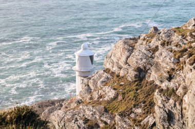 Uçurumun kenarında bir kayanın arkasına gizlenmiş beyaz bir deniz feneri, Koyun Başı, Batı Cork, İrlanda