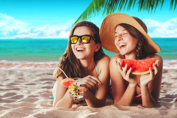 Vacation. Summer travel. Two girls young women in bikinis sunbathing on the tropical beach with cocktails and watermelon in sunglasses smiling at camera laying on the sand with palm and sky background — Stock Photo, Image