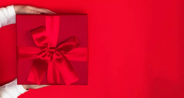 Hands baby holding gift for mom on March 8 and Mother\'s Day. Closed red box with a bow on a red background. The concept of gift giving.