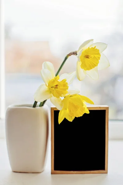 Three daffodils in a vase near an empty wooden frame with a black background to insert the image. Spring flowers on the windowsill.