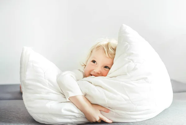 charming little blonde girl in a white bodysuit plays with a pillow on the bed at home. little charming girl lies on a white pillow. Cozy textiles for children