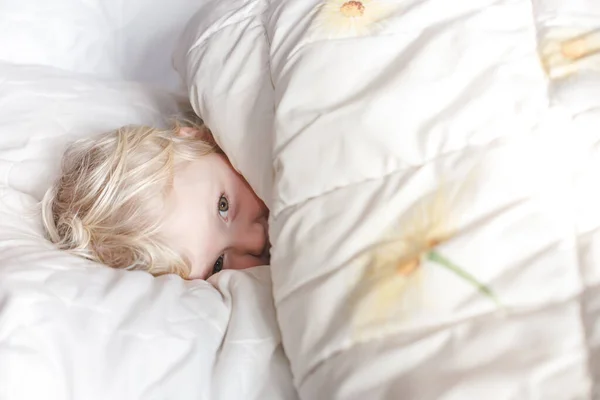 Carefree calm little girl lying on white bedding, hiding under a blanket, waking up in the morning from the sun\'s rays. The blonde slept in bed and opened her eyes cheerfully squinting. A naughty cheerful child.