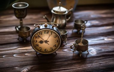 Tahta arka plandaki saat. Alarm çalıyor. Acele et! Acele et. Zaman doldu. Çok az zaman. Güneşin doğuşuyla aydınlanan bir masada gümüş mahsullü dama. İş günü için hazırlanılıyor.