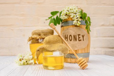 Taze akasya bal hasadı 2021 ahşap bir fıçıda ve üzerinde bez bir peçete olan şeffaf bir kavanoz. Çiçek açan beyaz akasya dalları, bal için çiseleyicinin yanındaki kızılgerdanlıklar.