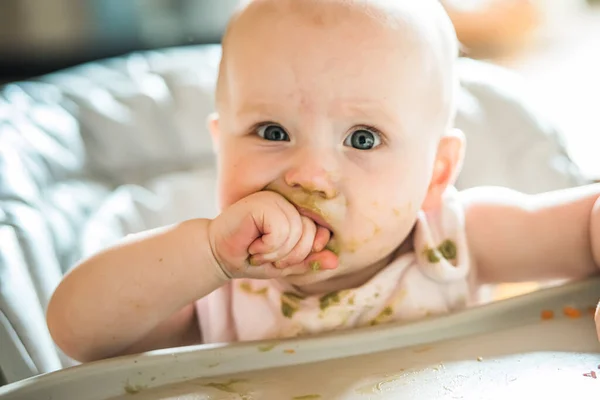 自宅でおいしい料理を食べるハイチェアで6ヶ月の赤ちゃんの女の子 面白いです赤ちゃんとともに土壌健康ブロッコリーPuree顔置く拳で口 — ストック写真