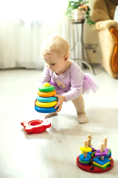 girl plays logical pyramid on floor in living room on sunny day. Montessori wooden toy folded pyramid. Circle, quadra, triangle, rectangle wooden elements of children\'s toys. Multi-colored toy blue, yellow, red, green