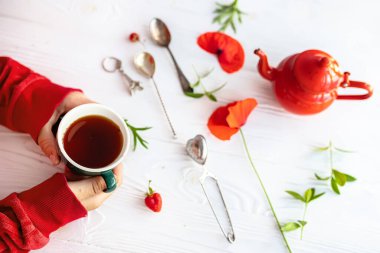 Hands holding a cup of coffee on background of white wooden table. Red teapot and fresh poppies during breakfast