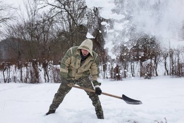 Kamuflajlı adam bahçedeki karları temizliyor. Ordu elinde kar küreği tutuyor.