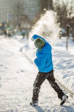 Mavi ceketli çocuk kar attı. Kartopu, çocuğu attıktan sonra birçok kar tanesine dağıldı. Kar güneşte parlıyor. Güneşli kış günü.