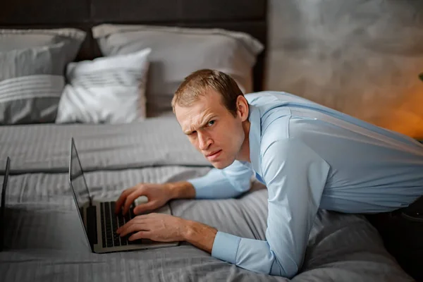 gloomy man raised eyebrow and looked indignantly. freelance businessman works on laptop sitting near bed in house. Confused businessman