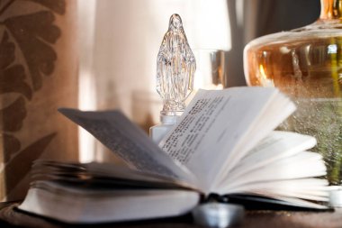 Bir Katolik İncili, yanında cam bir heykel olan bir masanın üzerinde açık duruyor. İçinde su olan cam bir vazo da var. Sahneye yumuşak bir ışık düşer ve sıcak bir atmosfer yaratır..