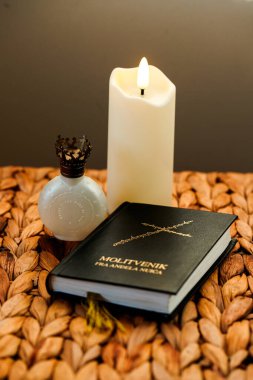 Masada bir mum, dekoratif bir şişe ve bir İncil var. Mum yanıyor, nesnelere ışık tutuyor. Örgü arka planda basit..