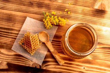 Bir bal peteği parçası bal kavanozunun yanındaki bir kağıt parçasına oturur. Tahta bir kaşık bal peteğinin yanında duruyor. Sarı çiçekler doğal bir dokunuş ekler. Sahne, bal eşyasının basit bir toplantısını gösteriyor..