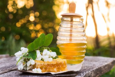 Bal peteği bal dolu bir kavanozun yanında bir tabakta oturur. Yeşil yapraklar ve beyaz çiçekler mevcuttur. Gündüz ortası doğal ışığı gösteriyor..