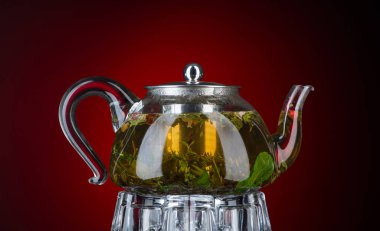 Kırmızı zemin üzerinde şeffaf bir çaydanlıkta naneli ve otlu çay. Arkaplan ışığıyla izole edilmiş