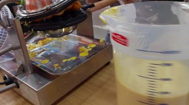 The process of making waffles at an ice cream shop. Yellow batter is poured into hot, round metal molds, creating a lively and appetizing kitchen atmosphere with a distinctive sweet aroma.