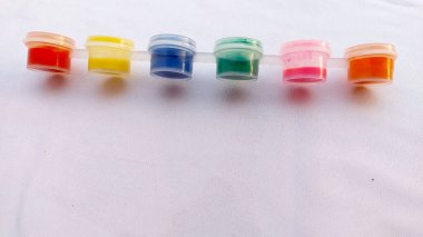 Renkli boyalardan oluşan küçük kaplar (kırmızı, sarı, mavi, turkuaz, pembe, turuncu) beyaz bir kumaş üzerinde beyaz kapaklar ile yatay olarak sıralanır..