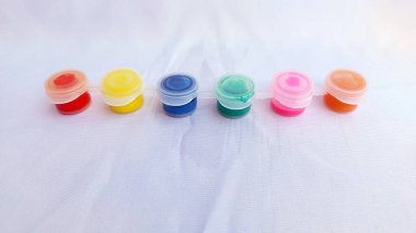 Renkli boyalardan oluşan küçük kaplar (kırmızı, sarı, mavi, turkuaz, pembe, turuncu) beyaz bir kumaş üzerinde beyaz kapaklar ile yatay olarak sıralanır..