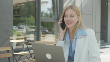 Gülümseyen İş Kadını Kafe 'de Smartphone ile konuşuyor. Modern Şehir Ayarları 'nda dizüstü bilgisayarla uzaktan çalışırken Kendine Güvenen Kadın Profesyonel Arkadaşça Telefon Sohbeti