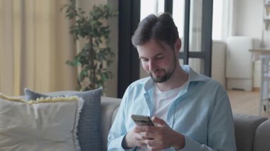 Parlak rahat oturma odasındaki koltukta Smartphone Content 'da gezinen adam. Günlük Tarayış, Sosyal Medya veya Haber Kaynağı Evdeki Huzurlu Dijital Anın tadını çıkarıyor. Yüksek kalite 4k görüntü