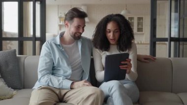Çift, sıcak oturma odasındaki koltukta tablet içeriğini tartışıyor. Koltuğa yakın oturan genç adam ve kadın dijital içeriği değerlendiriyor, konuşuyor ve parlak iç mekanda fikirleri paylaşıyorlar.