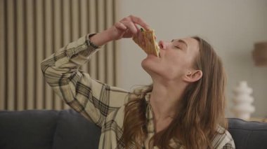 Kadınlar rahat ev içinde peynirli pizza dilimlerini seviyorlar. Aç Kadın Lezzetli Yemek, Tek Başına Yemek, Tatmin Olma, Kapalı Oturma Odasında Rahatlarken Tatmin Olma Arzusu