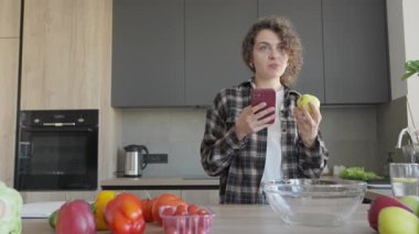 Modern ev mutfağında yemek pişirirken akıllı telefondan yemek tarifini kontrol eden bir kadın. Günlük Kıyafetlerde Odaklanan Kadın Parlak İç Kesimlerde Çevrimiçi Öğretim Kullanarak Sağlıklı Yemek Hazırlıyor