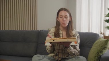 Evde rahat bir koltukta bacak bacak üstüne atarak oturan bir kadın pizza kutusunu tutuyor. Rahatlatılmış Modern Oturma Odası 'nda Rahatlık Yemeğinin Tadını Çıkartan Genç Kadın Konsepti