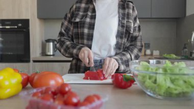 Sağlıklı ev yapımı salata için mutfak tezgahında taze sebzeleri dilimliyorum. Parlak modern mutfak iç kısmında ahşap yüzeyde çan biberlerini kesen ellere yakın, taze malzemelerle beslenen yemekler.