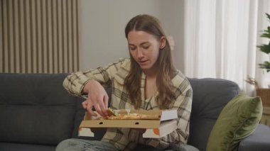Evde rahat koltukta dinlenirken pizzanın tadını çıkaran bir kadın. Rahat bir şekilde oturma odasında otururken lezzetli pizzalar yiyen genç bir kadınla sıradan atıştırma anı.