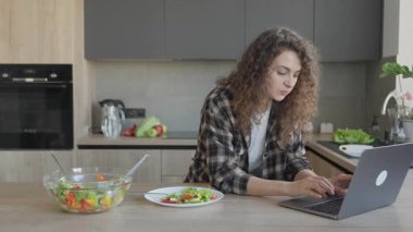 Modern mutfakta sağlıklı salata yerken dizüstü bilgisayarda çalışan bir kadın. Uzaktan Çalışma Konsepti, Sağlıklı Yaşam Tarzı, Dengeli Beslenme, Serbest Kadın, İçişleri Bakanlığı, Organik Gıda, Doğal Işık