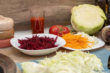 Vibrant, fresh chopped cabbage, grated beetroot, carrots, and diced onions are neatly arranged on a counter. A whole tomato, half cabbage, and tomato juice complete the scene, ready for a meal.