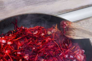 A close-up shot of vibrant shredded beets, carrots, and onions being sauted in a black frying pan, with steam gently rising. A wooden spoon stirs the healthy, colorful mixture.