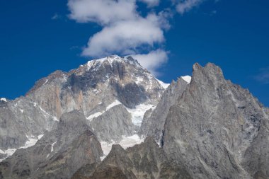 Derin mavi gökyüzünün altında dramatik kayalık dağ zirvesi ve dağınık beyaz bulutlar.