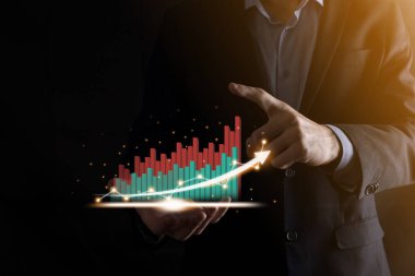 İş adamı, karanlık arka planda okları olan, istatistik, grafik ve grafiklerin büyüyen sanal bir hologramını tutuyor ve gösteriyor. Borsa. İş büyümesi, planlama ve strateji kavramı