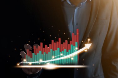 İş adamı, karanlık arka planda okları olan, istatistik, grafik ve grafiklerin büyüyen sanal bir hologramını tutuyor ve gösteriyor. Borsa. İş büyümesi, planlama ve strateji kavramı