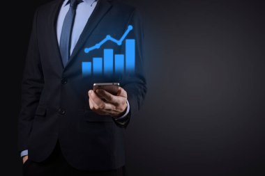 İş adamı elinde tablet tutuyor ve karanlık zemin üzerinde ok bulunan istatistik, grafik ve grafiklerin büyüyen sanal hologramını gösteriyor. Borsa. İş büyümesi, planlama ve strateji kavramı