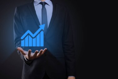 İş adamı elinde tablet tutuyor ve karanlık zemin üzerinde ok bulunan istatistik, grafik ve grafiklerin büyüyen sanal hologramını gösteriyor. Borsa. İş büyümesi, planlama ve strateji kavramı