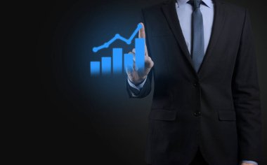 İş adamı elinde tablet tutuyor ve karanlık zemin üzerinde ok bulunan istatistik, grafik ve grafiklerin büyüyen sanal hologramını gösteriyor. Borsa. İş büyümesi, planlama ve strateji kavramı